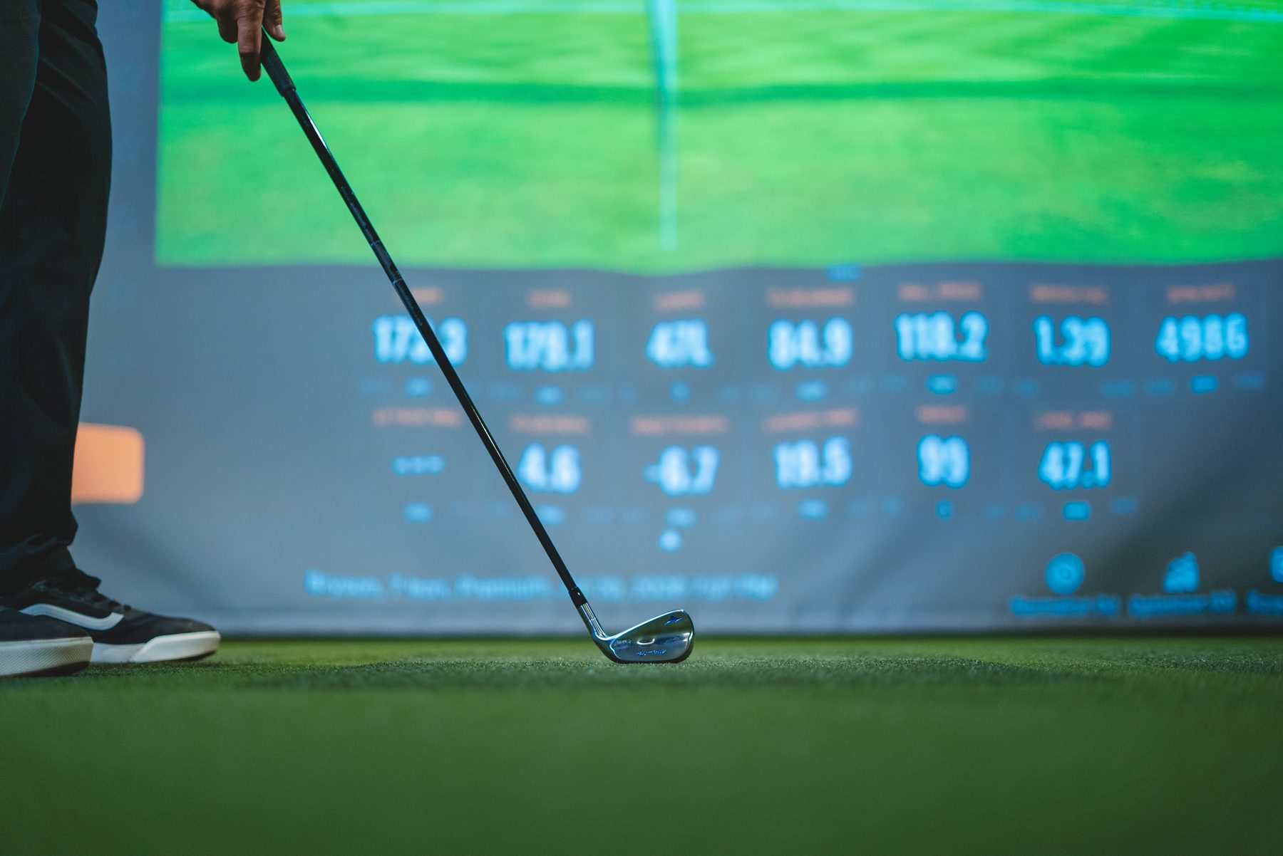 An iron at address with live TrackMan shot data on the bay screen