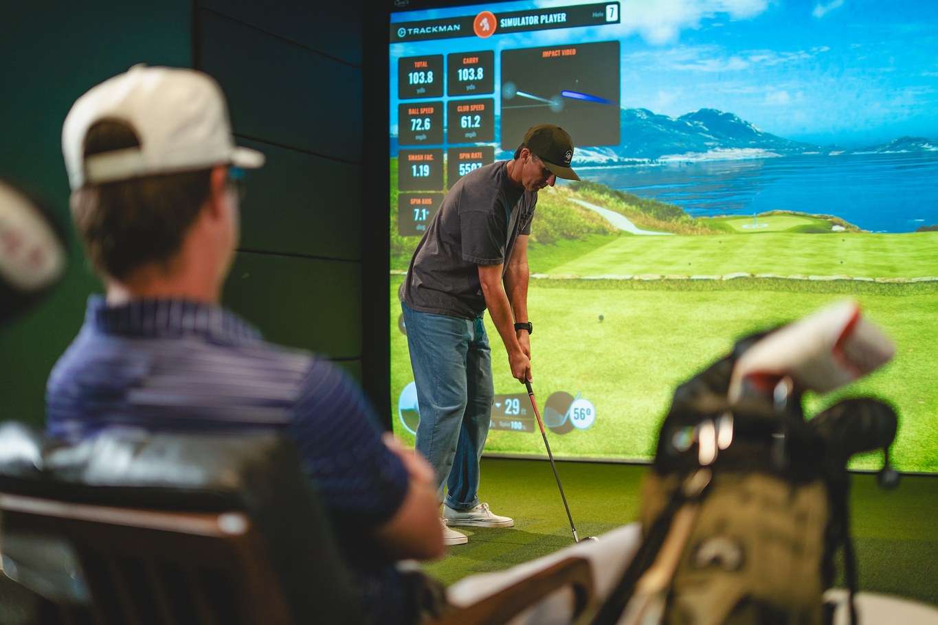 A golfer hitting into the simulator screen while a friend watches from the lounge seating